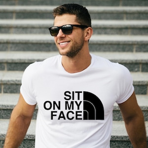 May include: White t-shirt with the text "SIT ON MY FACE" in black, with a black curved design. The shirt is being worn by a person wearing black sunglasses.