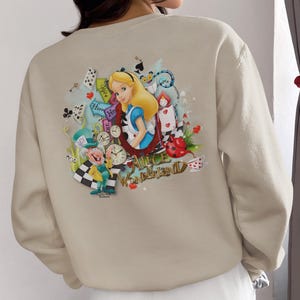 May include: Beige sweatshirt with a vibrant Alice in Wonderland design. The illustration includes Alice, the Mad Hatter, playing cards, clocks, and the words "Alice in Wonderland". The sweatshirt is a neutral colour.