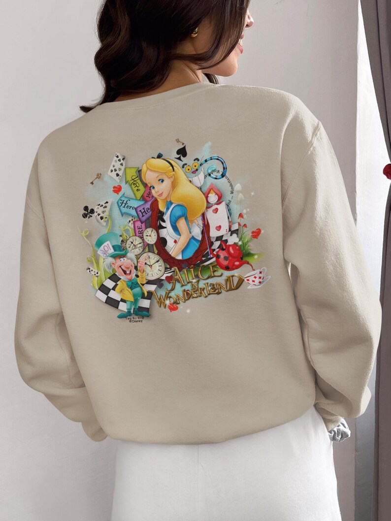 May include: Beige sweatshirt with a vibrant Alice in Wonderland design. The illustration includes Alice, the Mad Hatter, playing cards, clocks, and the words "Alice in Wonderland". The sweatshirt is a neutral colour.