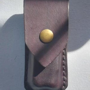 Könnte beinhalten: Eine dunkelbraune Lederscheide mit einem messingfarbenen Druckknopfverschluss. Die Scheide ist rechteckig mit einer Klappe, die sich umklappt und mit einem kreisförmigen Druckknopf befestigt wird. Die Kanten sind mit einem Kontrastfaden genäht.