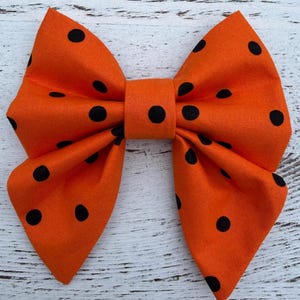 May include: A bright orange fabric bow featuring black polka dots. The bow is tied in the centre and has two large loops on each side. The polka dots are evenly spaced across the orange fabric.