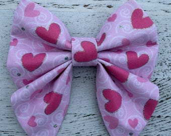 May include: A fabric hair bow featuring a light pink background with a pattern of dark pink hearts and subtle white swirls. The bow has a central knot and two large, folded loops on each side, creating a voluminous, symmetrical shape.