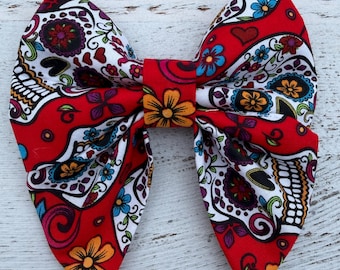 May include: A vibrant red fabric bow featuring a sugar skull design. The bow is adorned with colorful floral and skull patterns in white, blue, yellow, and purple. The bow is tied in the center with a matching red fabric.