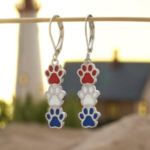 May include: Silver-tone earrings featuring a stacked paw print design in red, white, and blue. The earrings have a lever-back closure and are displayed against a blurred background of a lighthouse and beach.