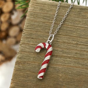 May include: A silver-toned metal necklace featuring a candy cane pendant. The pendant is decorated with diagonal red and white enamel stripes. The chain is a simple link style, attached to the top of the candy cane charm with a small jump ring.