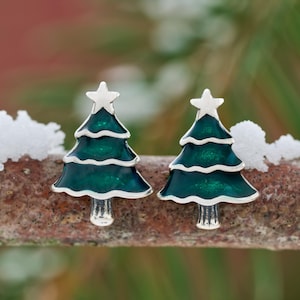 May include: A pair of Christmas tree stud earrings. Each earring features a silver-toned outline with a dark green enamel filling and a white star on top. The earrings are set against a blurred, natural background.