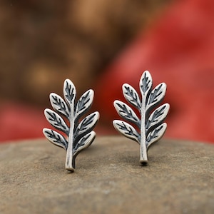 May include: A pair of silver-toned leaf stud earrings. Each earring features a detailed leaf design with multiple small leaves branching out from a central stem. The earrings are displayed on a stone surface against a blurred background.