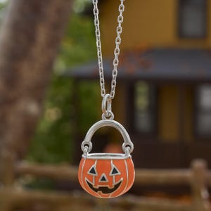 May include: A silver-toned necklace featuring a pendant shaped like a jack-o'-lantern bucket. The pendant has a bright orange enamel finish with black detailing for the eyes, nose, and mouth, suspended from a simple silver-toned metal chain link.