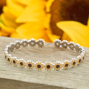 May include: Silver-toned bracelet featuring a repeating sunflower design. Each sunflower has yellow petals and a brown center. The bracelet is set against a wooden surface with a blurred background of yellow sunflowers.
