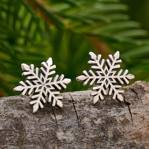 May include: A pair of silver snowflake stud earrings. Each earring is a detailed, symmetrical design with six points. The earrings are displayed on a piece of wood, with a blurred green background of foliage.