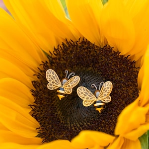 May include: A pair of bee-shaped earrings with gold-colored wings and black and yellow striped bodies, set against a bright yellow sunflower. The earrings are detailed with delicate wing patterns and antennae.