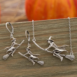 May include: A silver-toned jewelry set featuring a witch riding a broom. The set includes a pair of earrings and a necklace. The witch is wearing a pointed hat and flowing robes. The background is a wooden surface with an out-of-focus orange pumpkin.