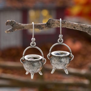 May include: A pair of silver-toned drop earrings shaped like miniature three-legged cauldrons. Each cauldron features a textured, pitted surface, a fixed handle, and a hook-style ear wire. Small, dark, rounded objects are visible inside each pot.