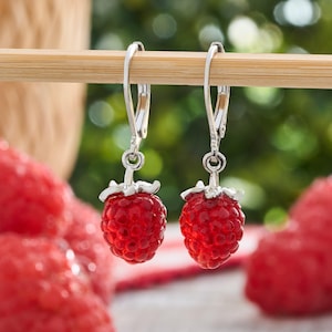 May include: A pair of dangle earrings featuring realistic red raspberry charms suspended from silver-toned leverback hooks. The berries have a textured, bumpy surface and a translucent appearance, hanging vertically from the metal findings.