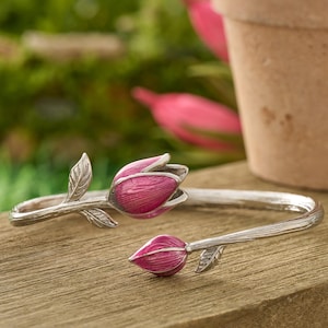 May include: A silver-toned bracelet featuring a tulip design. The bracelet has two pink tulip flower heads with green leaves. The bracelet is resting on a wooden surface with a blurred background of greenery and a terracotta pot.