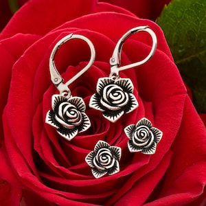 May include: Silver-toned rose earrings and two matching rose studs rest on a vibrant red rose. The earrings have a lever-back closure. The rose design features detailed petals and a spiral center, creating a classic floral theme.