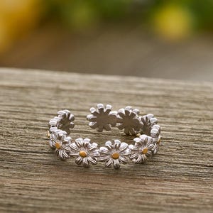May include: A silver daisy ring with yellow centers. The ring features a band of small, detailed daisy flowers. The ring is set on a wooden surface, with a blurred background of green and yellow.