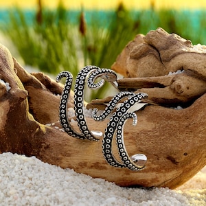May include: A pair of silver-toned hoop earrings shaped like octopus tentacles. The earrings feature a curved design with a textured surface covered in small, raised circular suction cups, creating a detailed, realistic appearance for the jewelry.