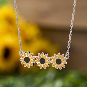 May include: A silver necklace featuring three sunflower pendants. Each pendant has a black center, surrounded by orange petals and a silver outline. The chain is delicate and the background is blurred.
