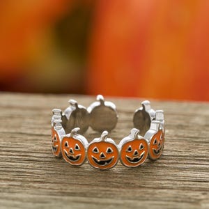 May include: A silver ring featuring a repeating pattern of orange jack-o'-lantern faces with black smiles and triangular eyes. The ring is set against a blurred wooden surface and a warm orange background, perfect for Halloween.