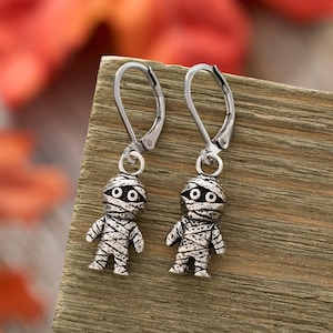 May include: A pair of silver-toned leverback earrings featuring small, three-dimensional mummy charms. Each charm depicts a figure wrapped in bandages with two circular eyes, crafted with an oxidized finish to highlight the textured details.