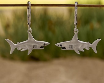 May include: A pair of silver-colored shark earrings. Each earring features a detailed shark design with fins, gills, and a toothed mouth. The earrings are suspended from silver lever-back closures, perfect for ocean lovers.