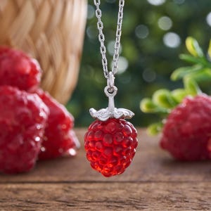 May include: A silver necklace featuring a pendant shaped like a realistic red raspberry. The pendant has a textured, bumpy surface and a silver stem cap at the top, suspended from a delicate silver chain link necklace.