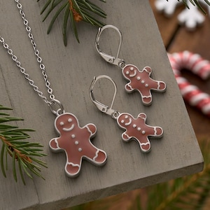 May include: A silver-toned jewelry set featuring a gingerbread man pendant necklace and matching earrings. The gingerbread men are light brown with white accents and a smiling face. The set is displayed on a gray surface with pine needles.