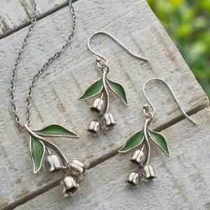 May include: A silver necklace and earrings set featuring a cluster of lily of the valley flowers with green enamel leaves.