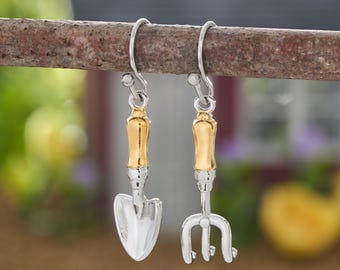 May include: A pair of dangle earrings featuring miniature gardening tools. One earring is a silver-toned trowel and the other is a silver-toned garden fork, both with gold-toned handles and silver-toned hooks for attachment to the ear.
