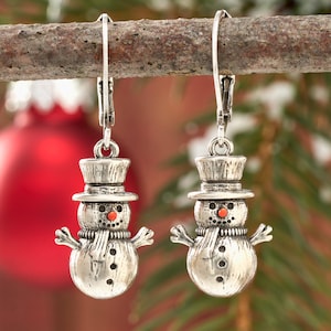 May include: Silver snowman earrings with a top hat, scarf, and orange nose. The earrings are hanging from a branch with a red ornament in the background. The snowman has black dot eyes and a button design.