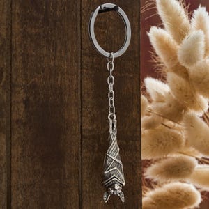 May include: A silver-colored bat keychain hanging against a dark wood background. The bat is detailed with etched wings and a small face. The keychain includes a chain and a circular ring.