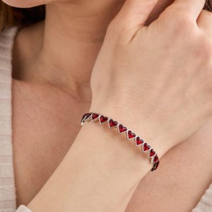 May include: A silver bracelet featuring a series of red heart-shaped charms. The bracelet is worn on a wrist, showcasing the delicate design and vibrant color of the hearts. A piece of jewelry perfect for a romantic gift.