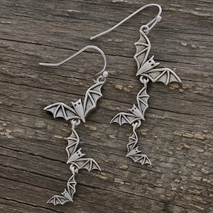 May include: A pair of silver-toned drop earrings featuring a vertical chain of three stylized bat figures. Each bat has detailed wing textures and is connected by small metal links. The earrings are attached to curved silver-toned ear wires.