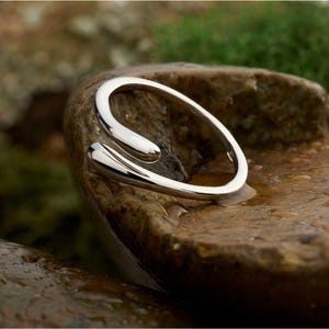 May include: A polished silver-toned open ring featuring a bypass design with two rounded, teardrop-shaped ends that curve toward each other. The metal surface is smooth and reflective, creating a continuous loop that remains open at the front.