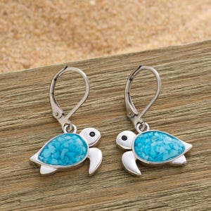 May include: A pair of silver-tone leverback earrings featuring turtle designs. The turtles have white bodies, black eyes, and turquoise-colored shell inlays. The earrings are displayed on a wooden surface.