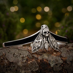 May include: A silver-toned metal cuff bracelet featuring a central moth-shaped ornament with a skull head and detailed wing patterns. The band is accented with a smooth, black inlay that extends along the sides of the cuff, creating a high-contrast look.