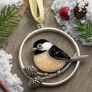 May include: A silver-toned bird ornament with a chickadee design. The bird has a black head, white cheeks, and a tan body. It is perched on a branch with two pinecones inside a circular frame. A yellow ribbon is attached for hanging.