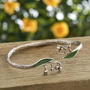 May include: A silver-toned bracelet with a textured, twig-like band. The bracelet features lily of the valley flower and leaf accents with green enamel detailing. The open cuff design is displayed on a wooden surface.