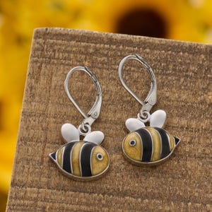 May include: A pair of silver-tone leverback earrings featuring a bee design. Each bee has a yellow and black striped body, white wings, and a small eye. The earrings are set against a wooden surface.