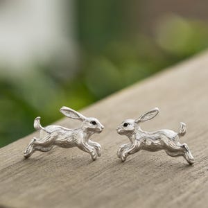 May include: A pair of silver-colored rabbit stud earrings. Each earring depicts a running rabbit with detailed fur texture and black eyes. The earrings are displayed on a wooden surface against a blurred green background.