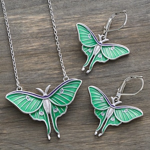 May include: A silver-toned jewelry set featuring a pendant necklace and a pair of earrings, all designed with a green and silver moth motif. The moth wings have a detailed, textured appearance with a purple accent. The earrings have a lever-back closure.