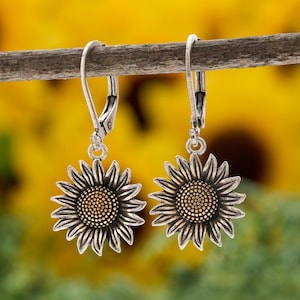 May include: A pair of silver-toned metal earrings featuring a sunflower design with a textured center and radiating petals. The earrings are attached to lever-back closures, showcasing a detailed metallic finish with darkened recessed areas.