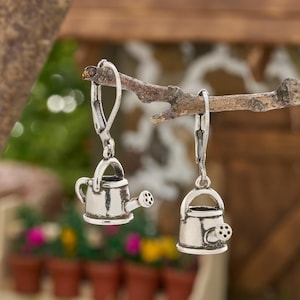 May include: Silver-toned earrings shaped like miniature watering cans, each with a handle, spout, and decorative details. The earrings are suspended from a branch against a blurred background of greenery and flower pots.