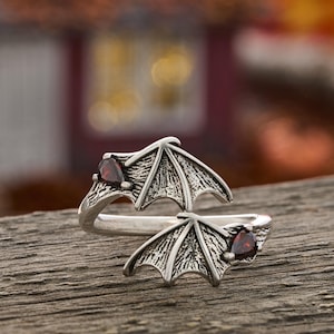 May include: A silver-toned open ring featuring two symmetrical bat wing designs. Each wing is accented with a small, dark red, pear-shaped gemstone set at the outer edge. The metal surface has a textured, oxidized finish throughout.