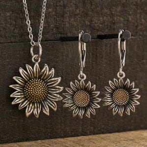 May include: A jewelry set consisting of a silver-toned sunflower pendant on a delicate chain and two matching sunflower drop earrings. Each sunflower features oxidized silver petals and a textured, golden-brown center, creating a detailed floral design.