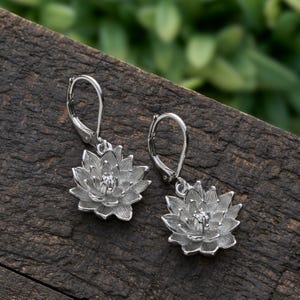 May include: A pair of silver-colored lotus flower earrings. Each earring features a detailed, open lotus blossom design with a small, central accent. The earrings are displayed on a dark, textured wooden surface, with a blurred green background.