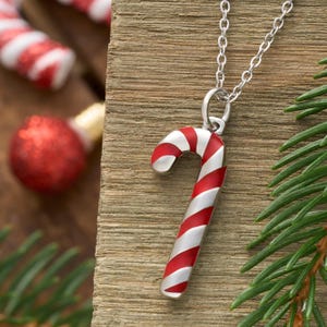 May include: A silver necklace with a candy cane pendant. The candy cane is red and white striped, with a curved shape. The necklace is displayed on a wooden surface, with Christmas ornaments in the background.