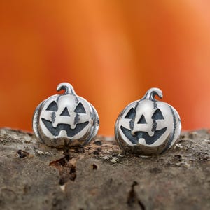 May include: Two silver-colored pumpkin earrings with carved faces, perfect for Halloween. Each earring has a stem and a smiling jack-o'-lantern face. The earrings are displayed on a piece of wood against an orange background.