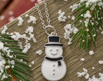 Puede incluir: Un collar con colgante de muñeco de nieve plateado. El colgante presenta un muñeco de nieve blanco con sombrero de copa negro, bufanda y tres botones negros. El collar se muestra sobre una superficie de madera con agujas de pino y nieve artificial.